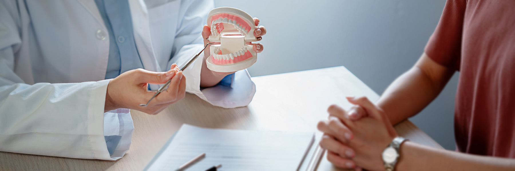 The image shows two individuals at what appears to be a dental office setting, with one person holding a model mouth open for examination while another person is seated behind a desk, possibly a dentist or dental hygienist, engaged in conversation with the model mouth holder.