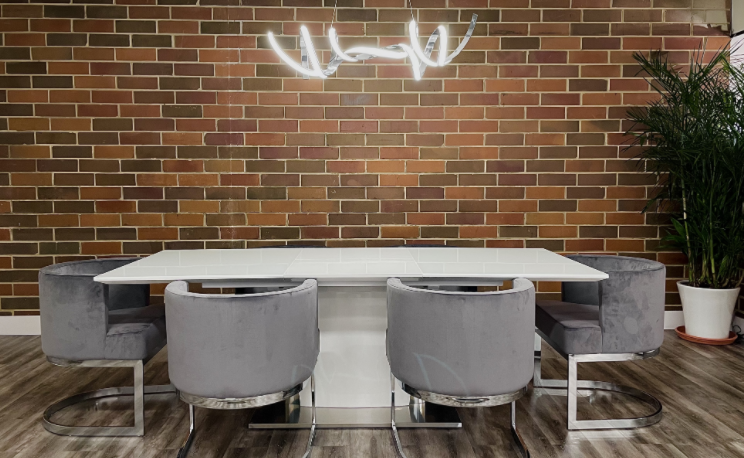 A modern office space with a brick wall, a white dining table surrounded by gray chairs, a large window with a view of the outside, and a contemporary design aesthetic.