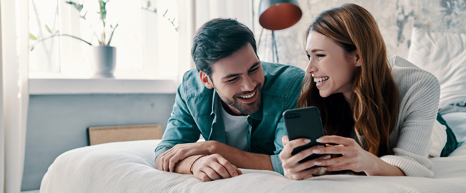 A young couple sharing a joyful moment while sitting on a bed with a smartphone between them.