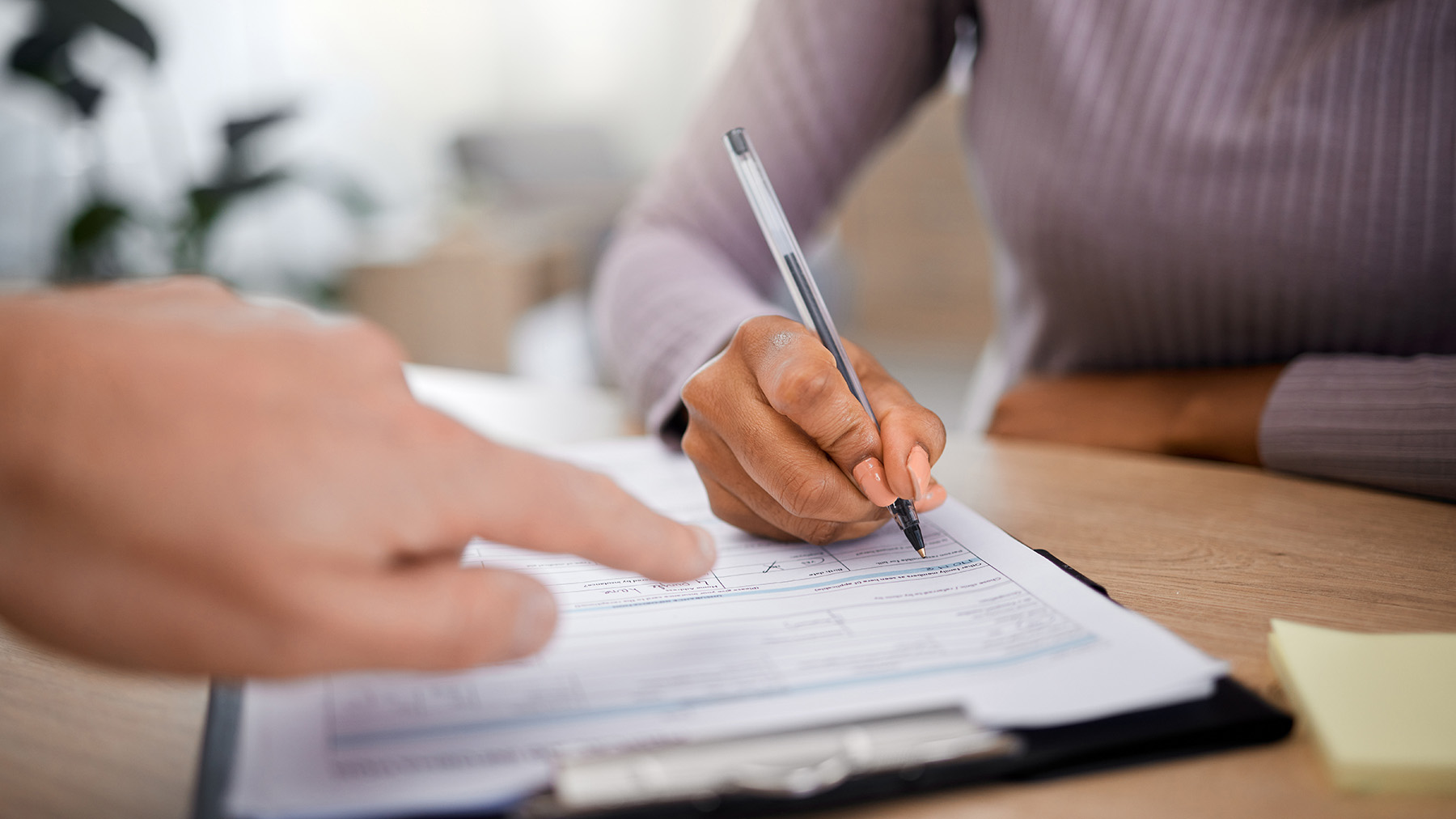 The image shows two individuals engaged in a professional activity at a desk with paperwork, including a clipboard and pen, indicating a business or office setting.
