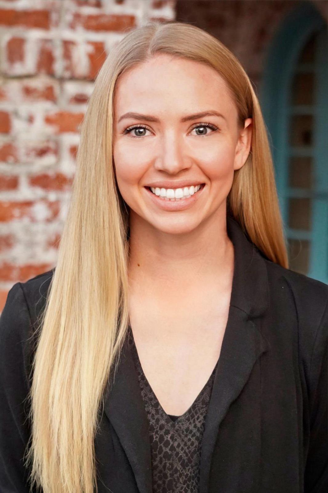 The image shows a woman with long blonde hair, smiling at the camera, wearing a black top and standing against a brick wall.