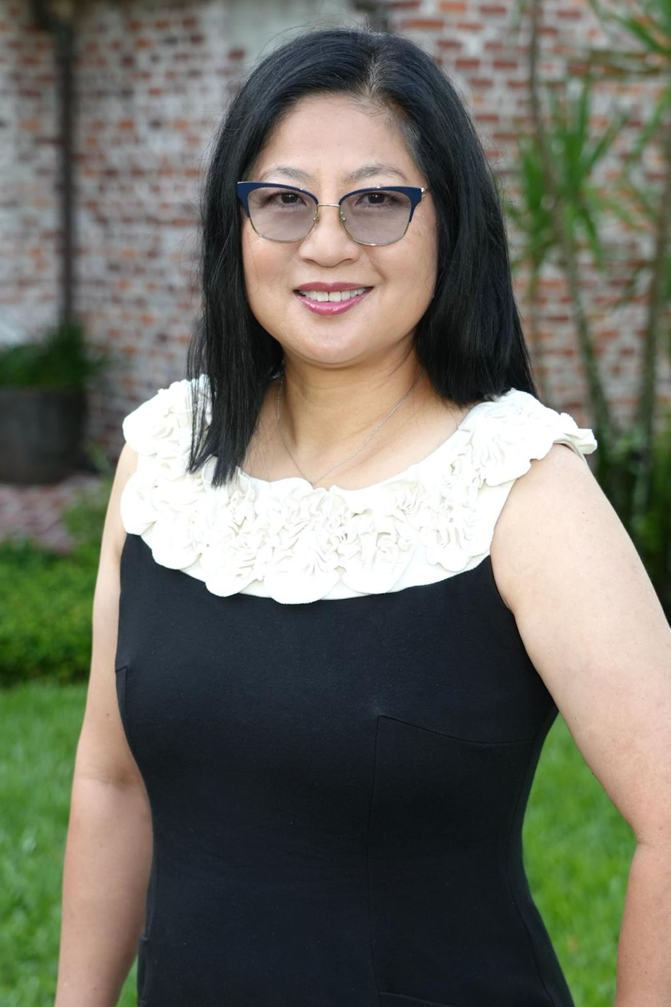 The image shows a person standing outdoors, wearing glasses, with dark hair and a black top, posing for a photo.